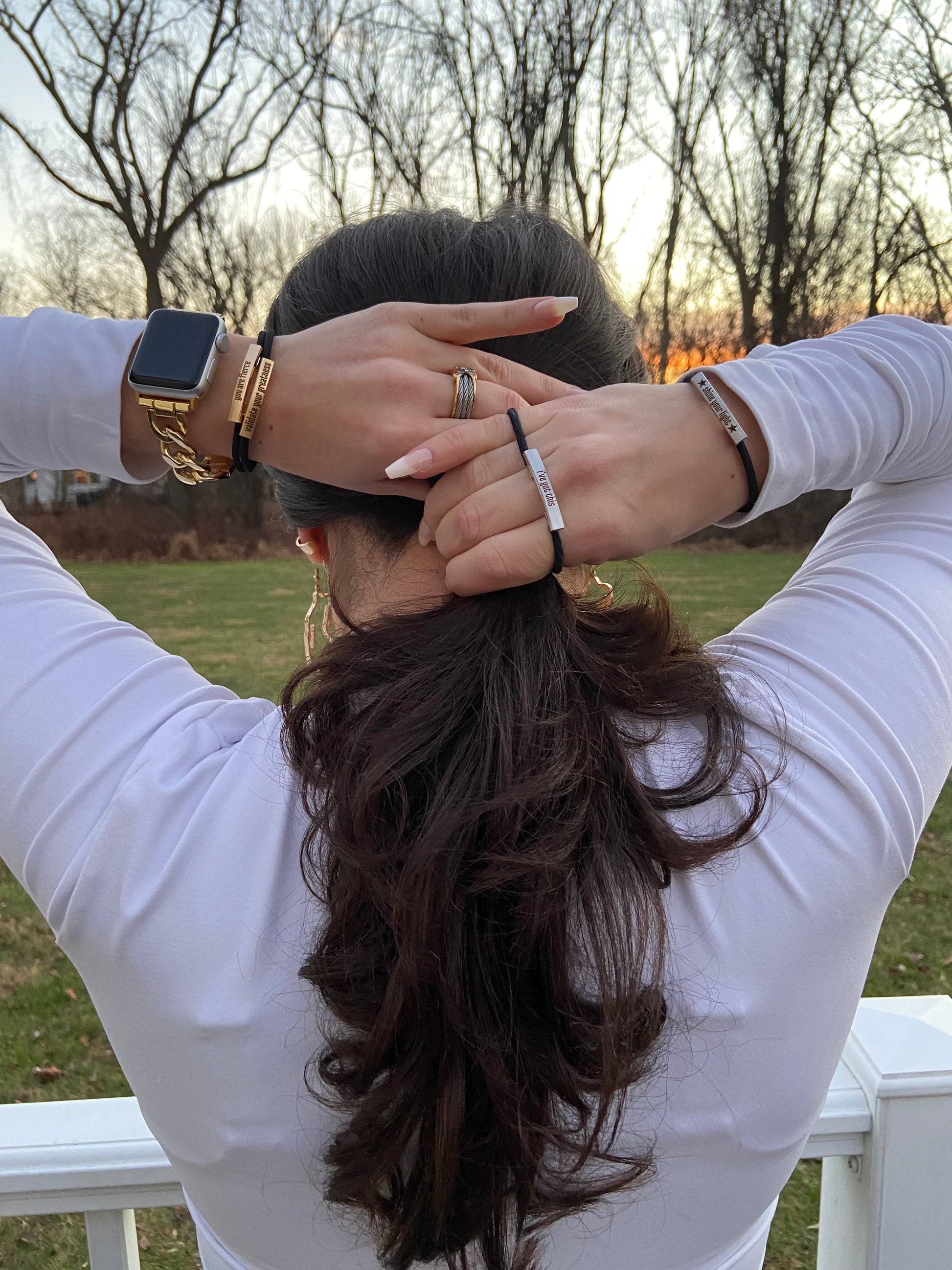 Woman putting her hair in a ponytail using bracelet~hair ties. She is wearing a white shirt and has long nails. Bracelet~hair ties are adorned on her wrists. The hair ties are made of black elastic with a gold or silver engraved bar.